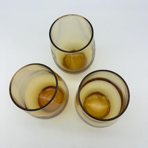 May include: Three amber-colored, stemless drinking glasses. The glasses have a rounded shape with a thick base and a dark brown rim. The interior of the glasses is a lighter amber color.
