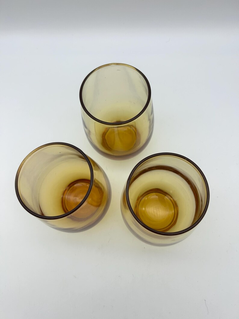 May include: Three amber-colored, stemless drinking glasses. The glasses have a rounded shape with a thick base and a dark brown rim. The interior of the glasses is a lighter amber color.