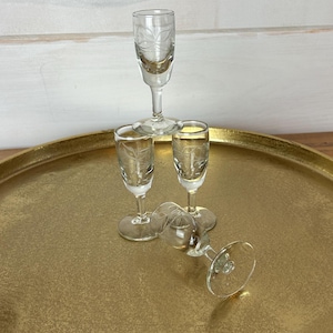 May include: Four clear glass shot glasses with etched designs on a gold tray. The glasses are stacked on top of each other, with one glass lying on its side.