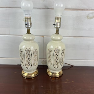 May include: A pair of white ceramic table lamps with gold accents and floral designs. The lamps have a classic style and would be a great addition to any home.