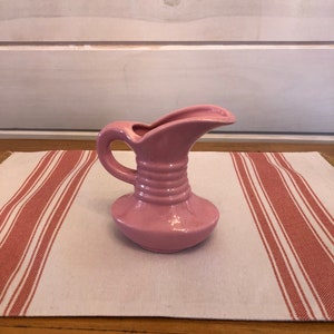 May include: A small pink ceramic pitcher with a handle and a spout. The pitcher is sitting on a white and red striped tablecloth.