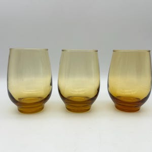 May include: Three amber-colored stemless glasses. The glasses have a rounded shape with a slightly flared base. The color transitions from a lighter amber at the top to a deeper amber at the base. The glasses are empty.