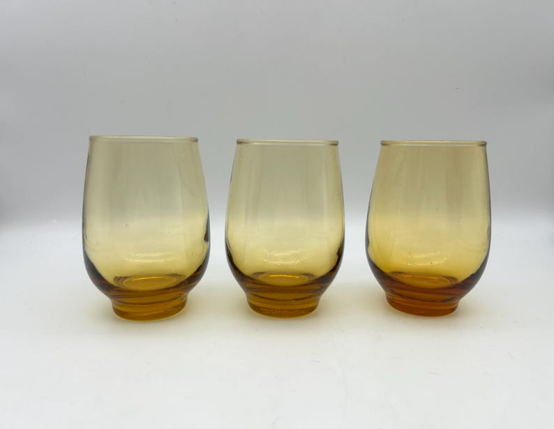 May include: Three amber-colored stemless glasses. The glasses have a rounded shape with a slightly flared base. The color transitions from a lighter amber at the top to a deeper amber at the base. The glasses are empty.