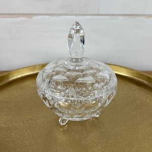 May include: A clear glass candy dish with a lid and three legs. The dish has a cut glass design.