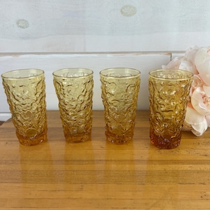 May include: Four amber-colored drinking glasses with a textured, bubbly pattern. The glasses are arranged in a row on a wooden surface, with a bouquet of pink flowers in the background. The glasses are approximately 6 inches tall.