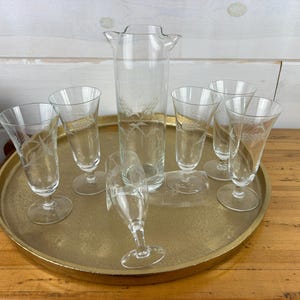 May include: A set of seven clear glass cocktail glasses with a wheat design. The glasses are arranged on a gold tray.