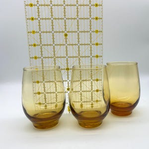 May include: Three amber-colored stemless glasses with a rounded shape. A clear ruler with yellow markings is behind the glasses. The glasses have a gradient color, with a darker amber base.