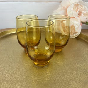 May include: Three amber-colored stemless glasses are arranged on a gold-toned tray. The glasses have a rounded base and a slightly flared rim. Soft pink flowers are visible in the background, adding a touch of color.