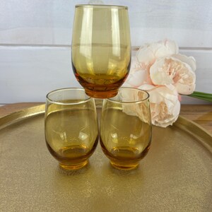 May include: Three amber-colored stemless drinking glasses. One glass is stacked on top of two others. The glasses are on a gold-colored tray. A bouquet of pink flowers is in the background.