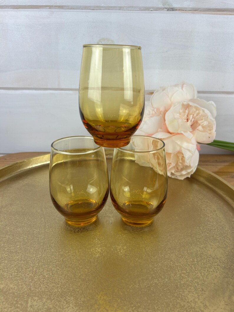 May include: Three amber-colored stemless drinking glasses. One glass is stacked on top of two others. The glasses are on a gold-colored tray. A bouquet of pink flowers is in the background.