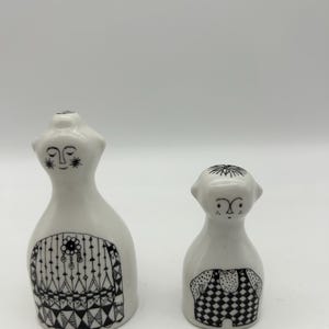 May include: A set of two white ceramic salt and pepper shakers with black and white geometric designs. The shakers are shaped like people with simple faces.