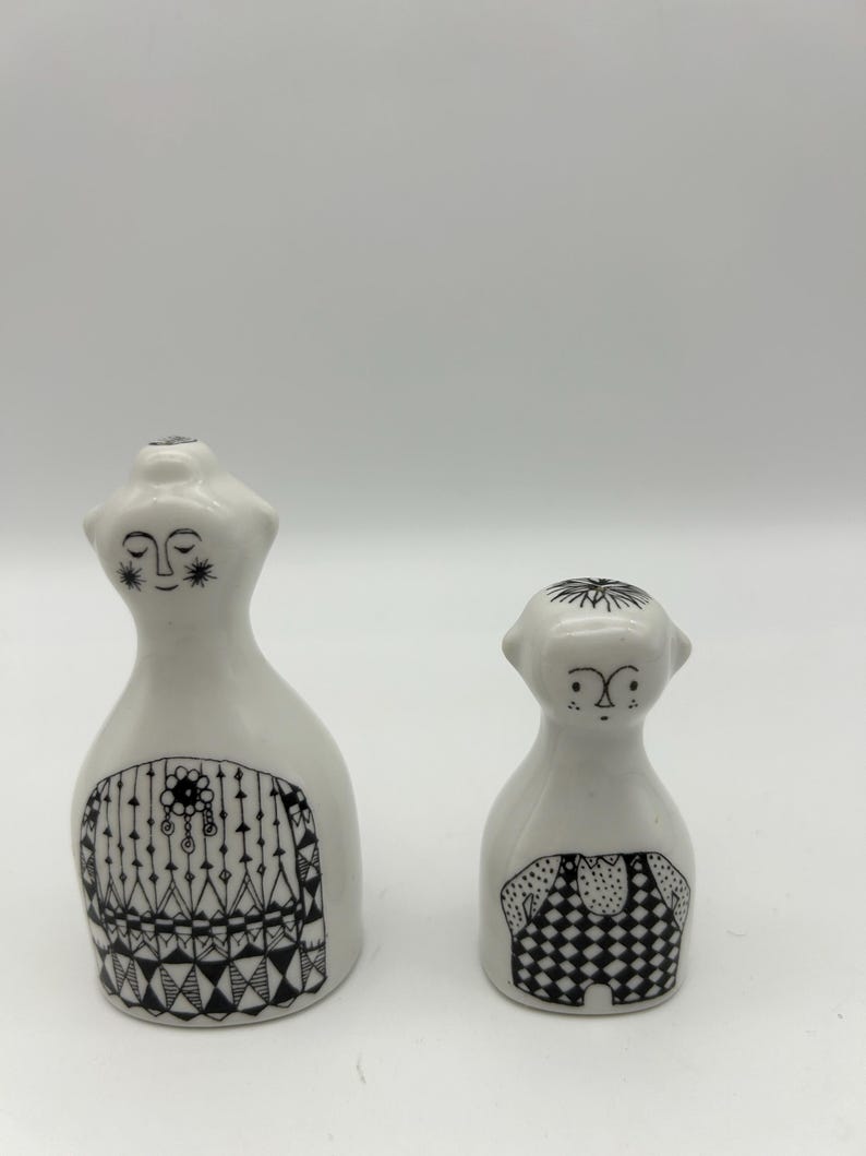 May include: A set of two white ceramic salt and pepper shakers with black and white geometric designs. The shakers are shaped like people with simple faces.