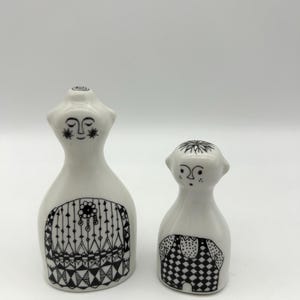 May include: A pair of ceramic salt and pepper shakers in the shape of a woman and a man. The woman is wearing a black and white patterned dress and the man is wearing black and white checkered pants.