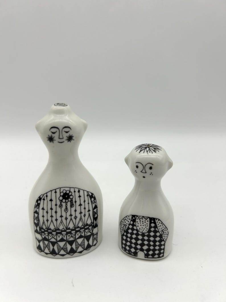 May include: A pair of ceramic salt and pepper shakers in the shape of a woman and a man. The woman is wearing a black and white patterned dress and the man is wearing black and white checkered pants.
