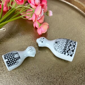 May include: A pair of white ceramic salt and pepper shakers with black and white geometric designs. The shakers are shaped like people and are standing on a gold tray.