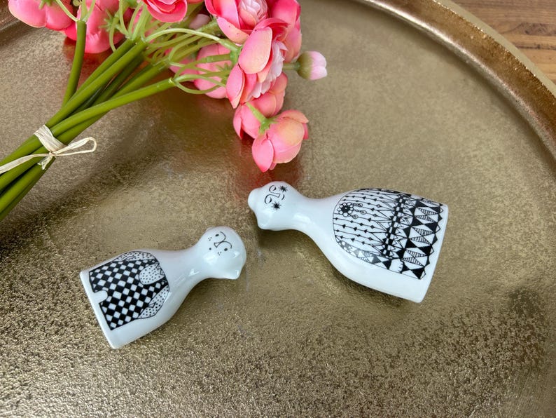 May include: A pair of white ceramic salt and pepper shakers with black and white geometric designs. The shakers are shaped like people and are standing on a gold tray.