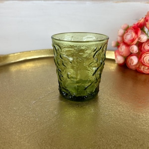 May include: A green glass tumbler with a textured, organic pattern. The glass is a vibrant shade of green and sits on a gold-colored tray. The background includes a white surface and a bouquet of pink flowers.