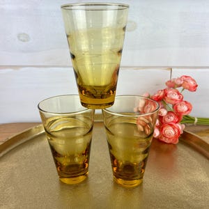 May include: Three amber-colored drinking glasses, one balanced atop two others, arranged on a gold-toned tray. The glasses have a classic, slightly flared shape. A bouquet of pink flowers is visible in the background.