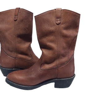 May include: A pair of brown leather cowboy boots. The boots have a mid-calf height, a rounded toe, and a stacked heel. The leather has a textured appearance. The soles are black.