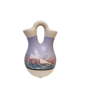 May include: A ceramic vase with a unique double-spout design. The vase features a light purple body with a geometric fish design in shades of blue, red, and white. The top and base are off-white. A decorative piece for the home.
