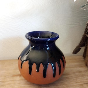 May include: A small, handcrafted ceramic vase with a rounded body and a dark blue interior. The vase features a terracotta base with black, dripped glaze detailing. The vase is suitable for small floral arrangements or decorative purposes.
