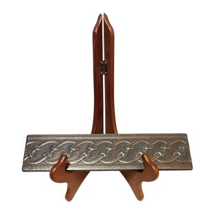 May include: A decorative silver-colored rectangular object with an embossed chain design, displayed on a wooden easel. The easel has a dark brown finish and a hinged back for stability. The object is approximately 12 inches long.