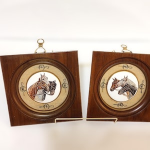 May include: Two framed horse portraits. Each features a circular image of two horses in a detailed painting. The frames are square with a dark wood finish and a gold-colored ring around the horse image. A metal loop is at the top for hanging.