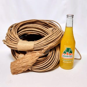 May include: A large, circular coil of light brown paper cord secured with a paper band, positioned next to a glass bottle of yellow Jarritos Passion Fruit soda with a silver cap and a label featuring a green pitcher graphic and text.