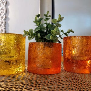 May include: Three textured glass candle holders in shades of yellow and orange. The center holder contains a small green plant. The holders are cylindrical and sit on a decorative gold surface.