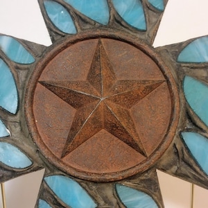 May include: A rustic cross with a central star design. The cross features a brown, weathered appearance with turquoise-colored glass inserts. The star is set within a circular frame, adding a decorative element to the religious symbol.