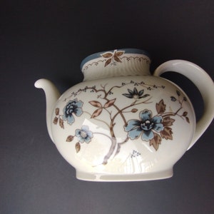 May include: Cream-colored ceramic teapot with a round body and a curved handle. The teapot is decorated with blue and brown floral patterns and a blue rim. The spout is curved and the lid has a small handle.