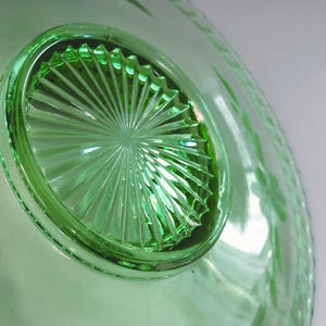 May include: A close-up of a green glass dish. The dish has a circular, textured base with a starburst design. The rim of the dish has a delicate floral pattern. The glass is translucent, allowing light to pass through.
