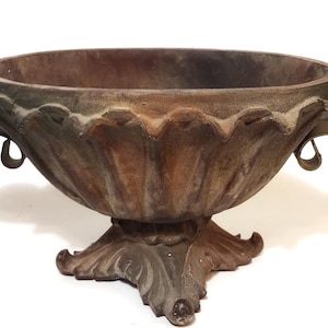 May include: A weathered, antique-style metal planter with a wide, scalloped rim and ornate handles. The planter has a pedestal base with decorative leaf accents. The overall color is a mix of brown and gray.