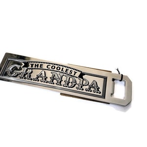 May include: Silver bottle opener with a black inscription that reads "The Coolest Grandpa".