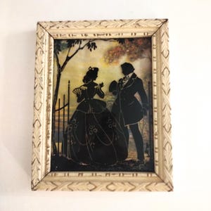 Puede incluir: Arte de silueta enmarcado que representa a una pareja con vestimenta de época. Las siluetas negras se sitúan sobre un fondo de paisaje, dentro de un marco crema ornamentado. La obra evoca una estética vintage.