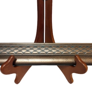 May include: A bronze-colored decorative trim piece with a scalloped pattern, displayed on a wooden easel. The trim has a metallic finish and is supported by a brown wooden stand, ideal for displaying art or decor.