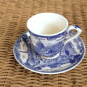 May include: A white teacup and saucer set with a blue and white illustration of the United States Capitol building. The cup has a curved handle and sits on a matching saucer with a similar design. The set is placed on a woven surface.