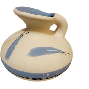 May include: A cream-colored ceramic pitcher with a rounded body and a curved spout. The pitcher features blue feather designs and a blue band around the base and rim. The handle has a circular opening.