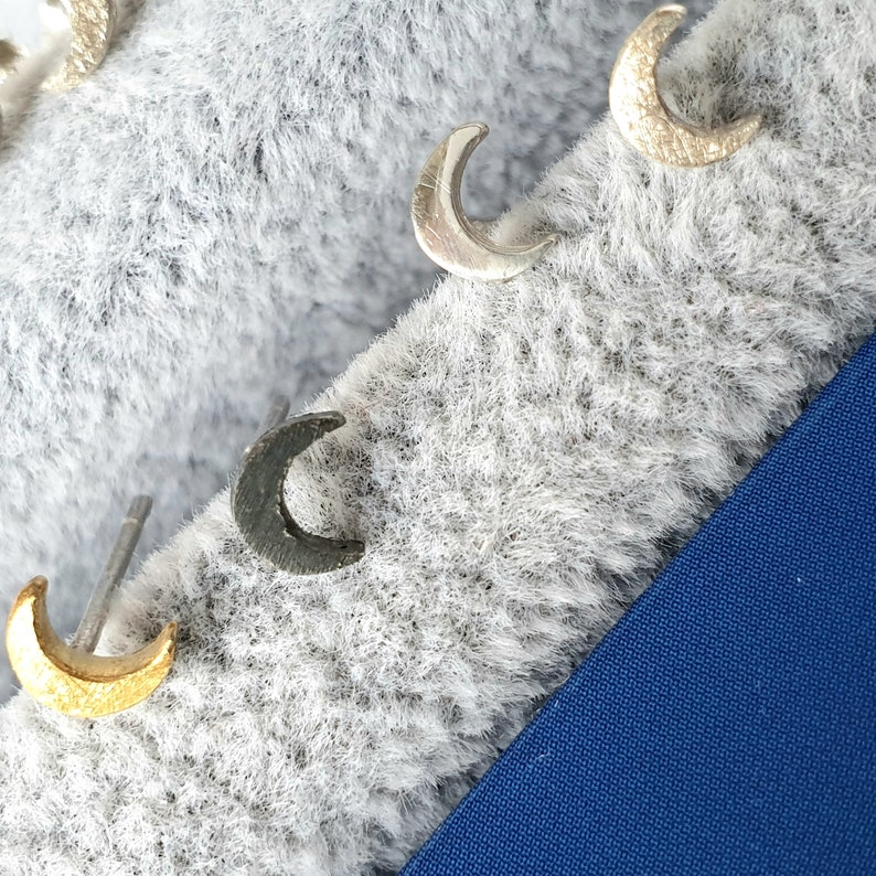 May include: Four silver crescent moon earrings on a white fuzzy surface. The earrings are in different shades of silver, black, and gold.