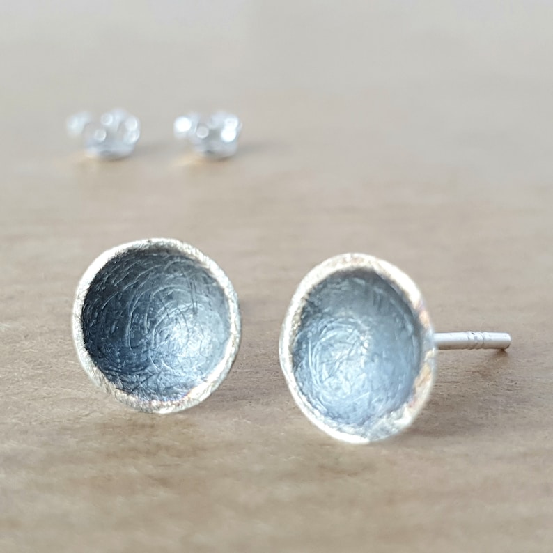 May include: A pair of silver stud earrings with a textured, circular design. The earrings have a slightly domed shape and a polished edge.