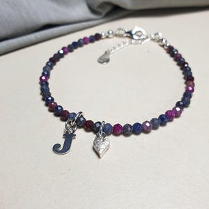 Könnte beinhalten: Ein Armband mit blauen, lila und roten Edelsteinen. Es hat einen Silberverschluss und zwei Anhänger: einen Buchstaben "J" und ein Herz. Das Armband liegt auf einer hellen Oberfläche.