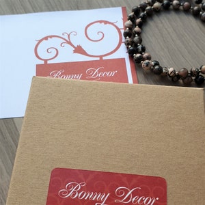 May include: A brown cardboard box with a red label that says "Bonny Decor" in a cursive font. The box is sitting on a wooden surface. A necklace of brown, black, and pink stones is lying on the surface next to the box.