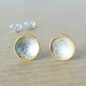 May include: A pair of silver and gold earrings with a round, textured design. The earrings have a gold rim and a silver center.