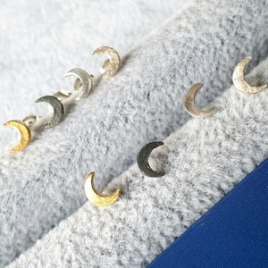 May include: A set of six crescent moon earrings in various finishes, including gold, silver, and black. The earrings are displayed on a gray fuzzy surface.