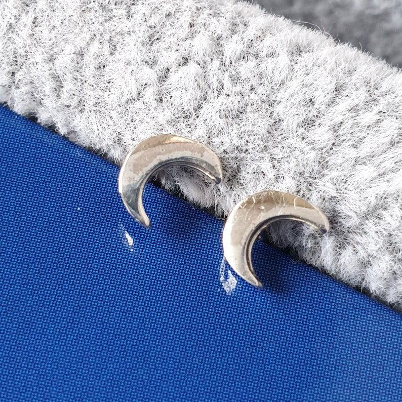 May include: A pair of silver crescent moon stud earrings. The earrings are small and delicate, and they have a simple, elegant design.