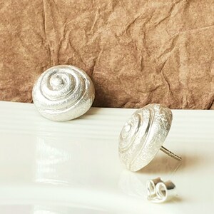 May include: A pair of silver earrings with a textured, spiral design. The earrings are in the shape of a shell.