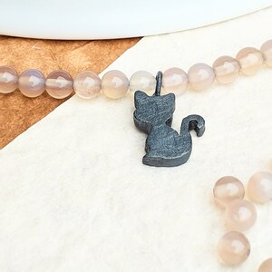 Könnte beinhalten: Ein schwarzer Katzenanhänger hängt an einer hellbraunen Perlenkette. Die Perlen sind aus Achat und haben eine natürliche, unregelmäßige Form.