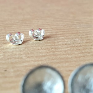 May include: Two silver earring backs, also known as butterfly backs, are shown on a wooden surface. The backs are designed to secure earrings to the earlobe.