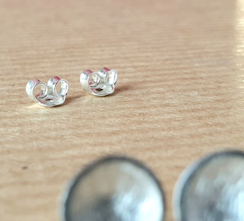 May include: Two silver earring backs, also known as butterfly backs, are shown on a wooden surface. The backs are designed to secure earrings to the earlobe.