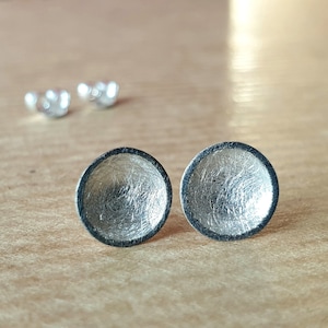 May include: A pair of silver earrings with a textured, circular design. The earrings have a dark gray rim.